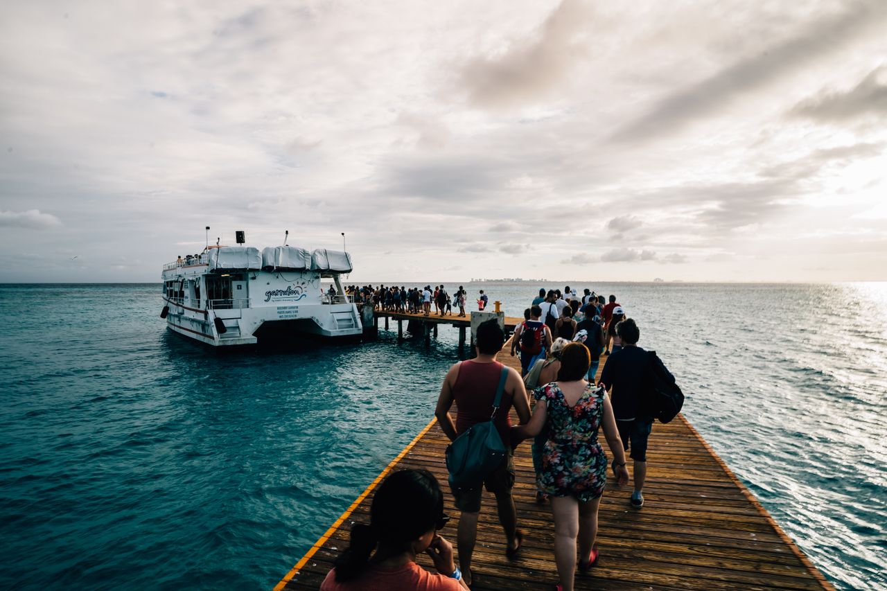 Tourists walk along a wooden pier to board a ferry under a dramatic sky, perfect for travel and vacation themes.